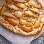 Close-up of a delicious homemade pear galette, topped with chopped nuts and rosemary, on a textured surface.